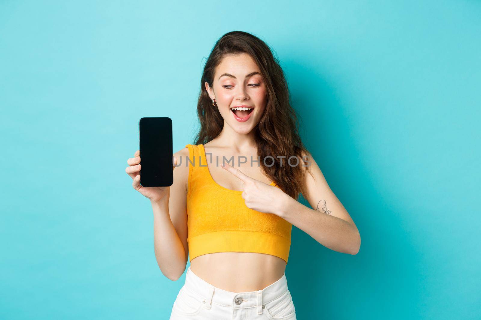 Young happy beautiful girl, pointing and looking at empty smartphone screen, demonstrate advertisement on mobile display, standing against blue background.