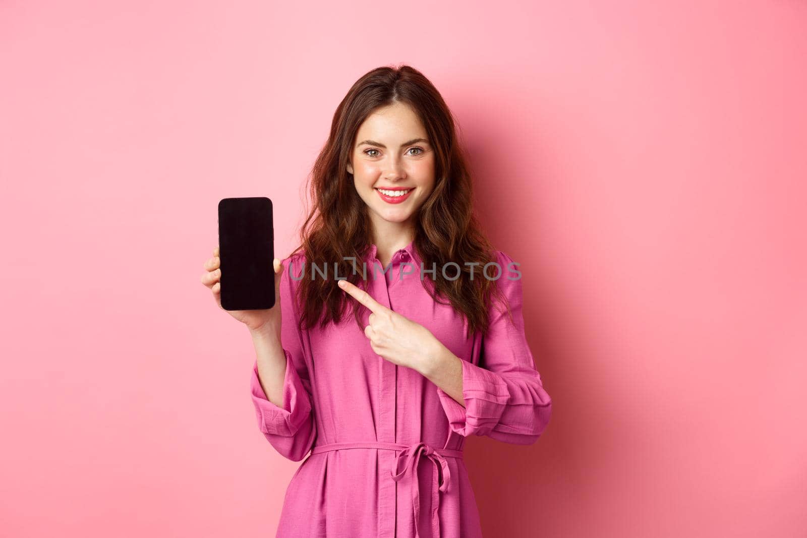 Young caucasian woman in stylish dress, pointing finger at smartphone screen and smiling, showing promo deal online, pink background.