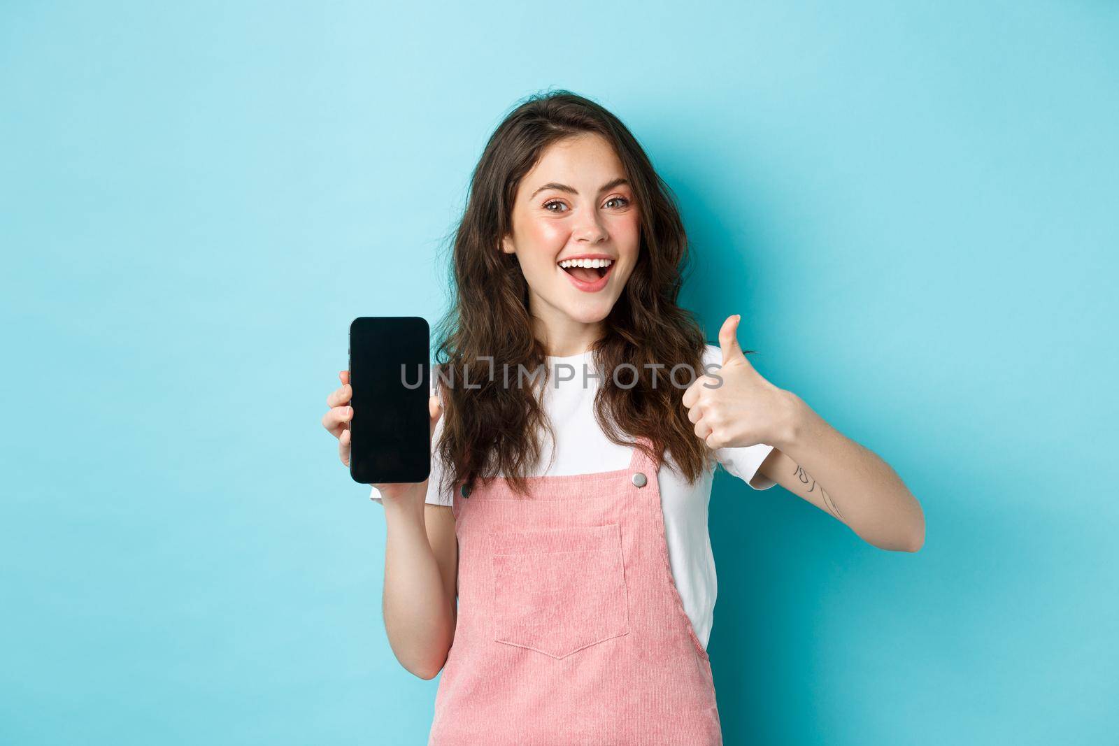 Yes this is good. Smiling cute girl showing thumb up and empty smartphone screen, recommending application or online store, standing over blue background by Benzoix