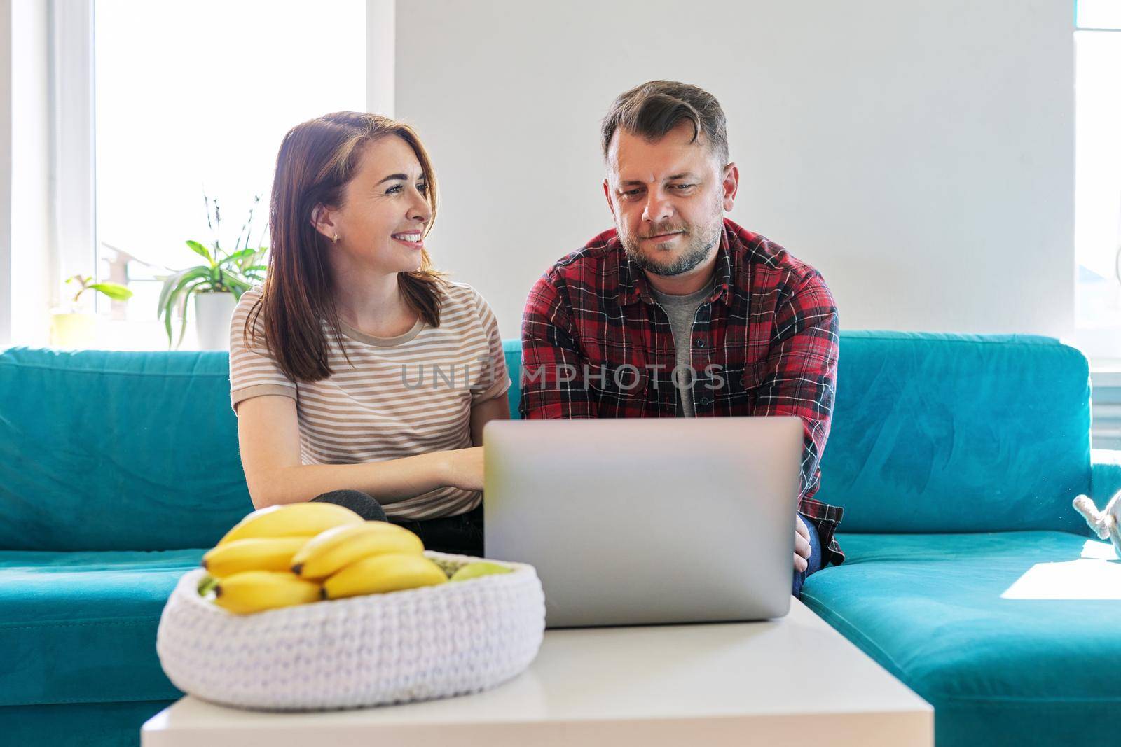 Middle aged couple sitting together at home on the couch looking into the laptop screen. Positive smiling husband and wife, lifestyle, relationship, technology concept