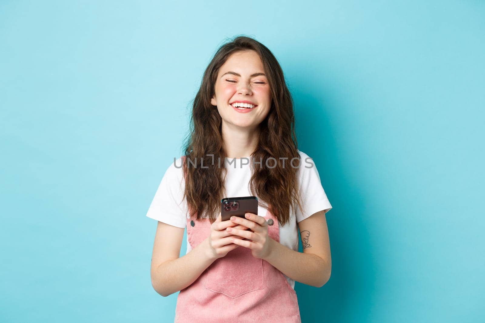 Portrait of happy young woman laughing with smartphone in hands, smiling and looking carefree, using mobile app, standing over blue background.