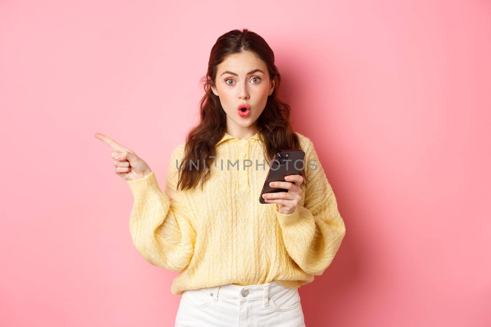 Surprised young beautiful girl gasping and saying wow, pointing finger aside at left copyspace, holding mobile phone, standing against pink background.