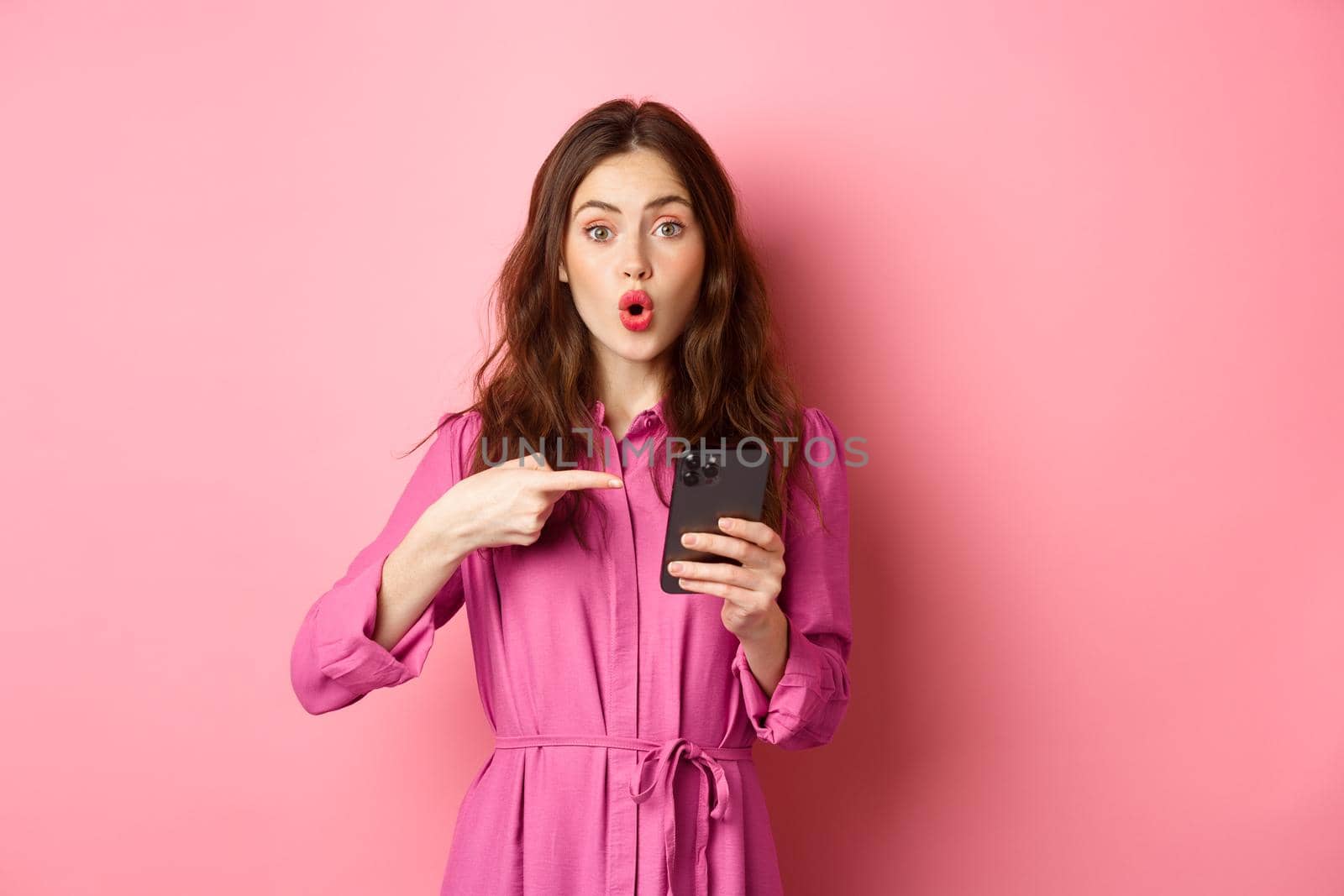 Young woman pointing finger at her phone and gasping wondered, talking about smartphone app, showing something interesting online, standing over pink background. Copy space