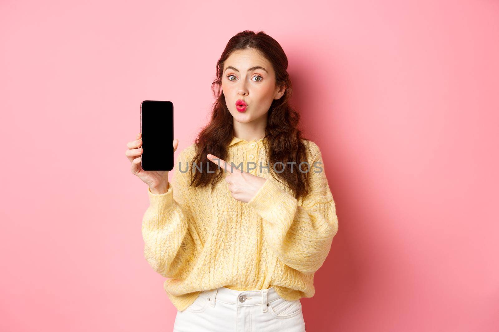 Technology and online shopping. Excited girl looks curious, points finger at empty mobile phone screen, shows app on smartphone, standing against pink background by Benzoix