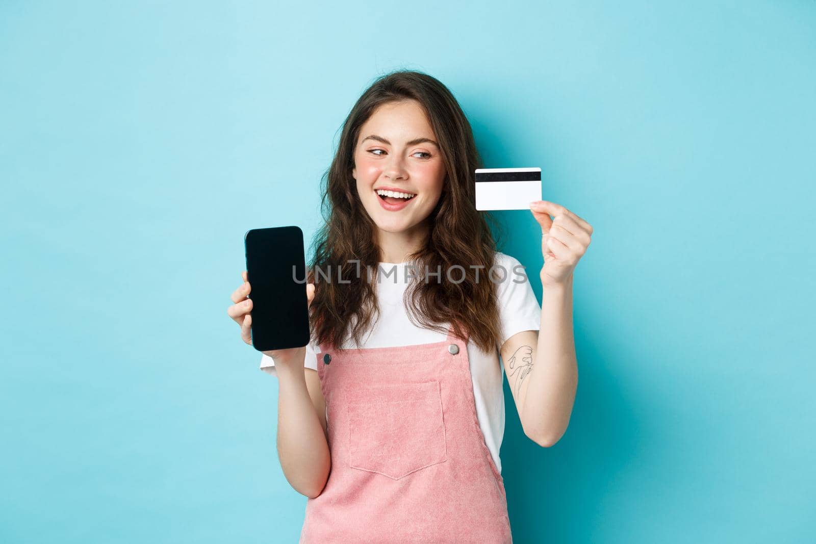Cheerful girl in summer clothes showing smartphone screen and plastic credit card, paying online, shopping, standing over blue background.