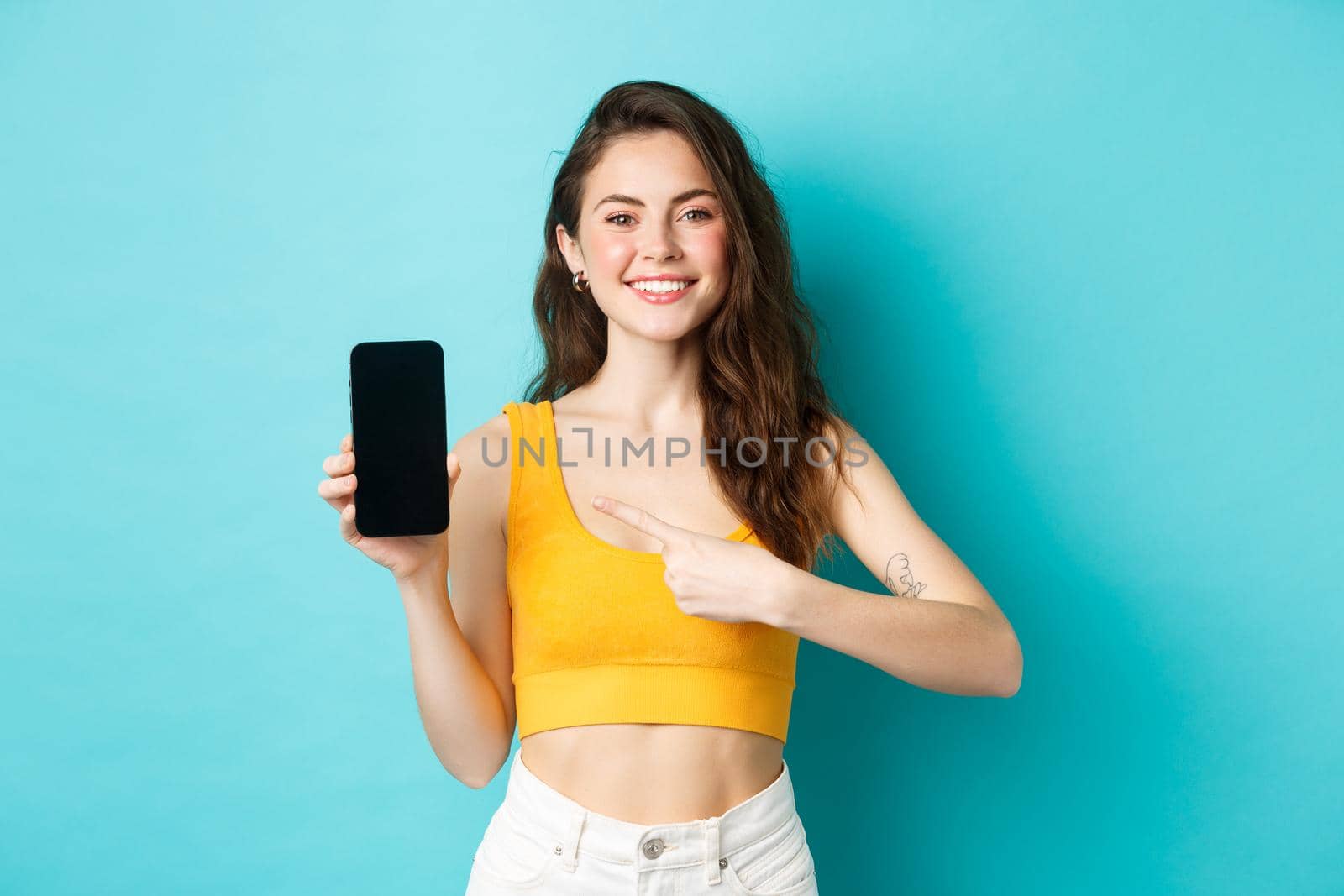 Happy attractive woman showing advertisement on smartphone screen, pointing at empty display of phone and smiling, standing over blue background.