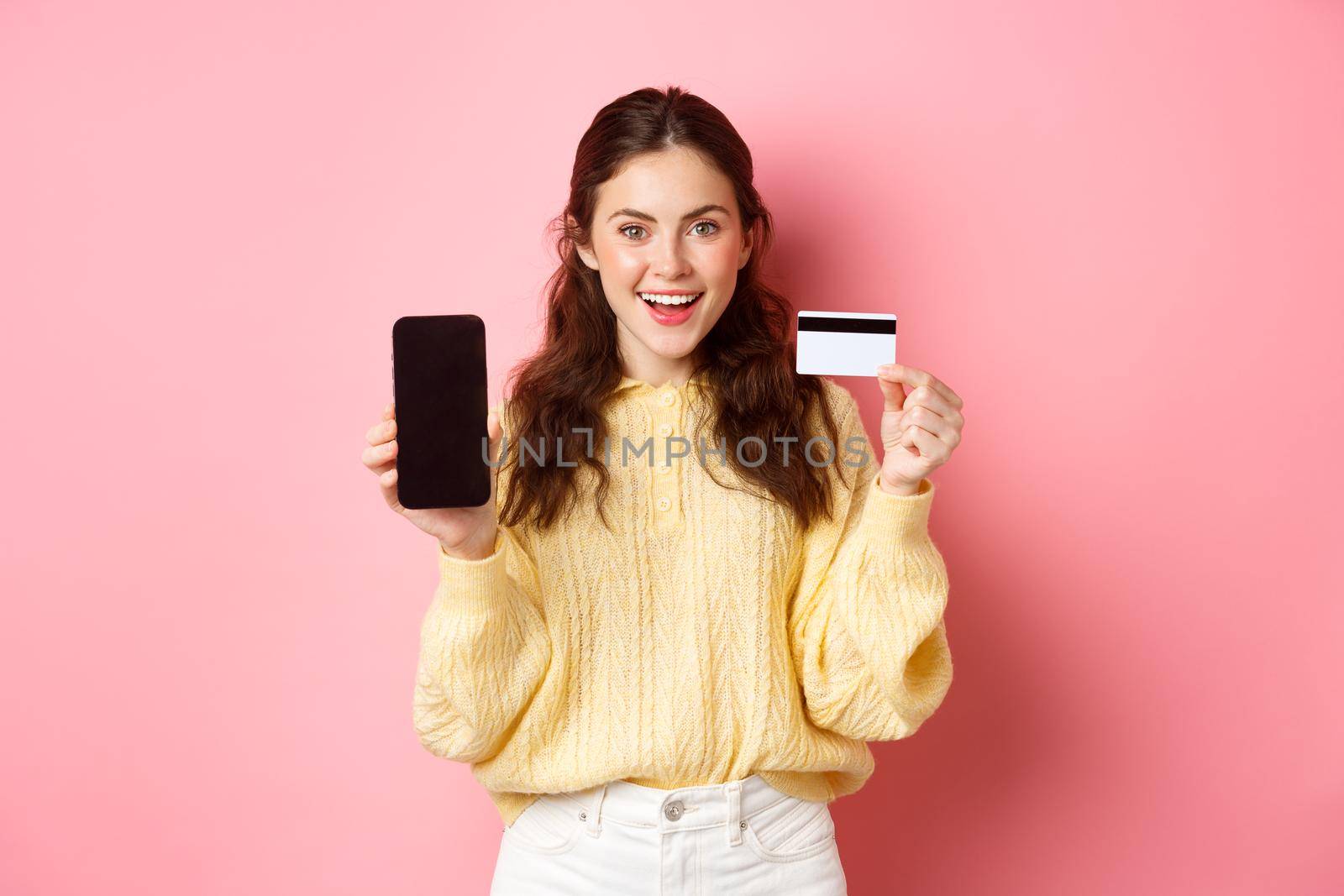 Technology and online shopping. Young attractive female model showing empty smartphone screen with plastic credit card, show bank account or application, standing over pink background.