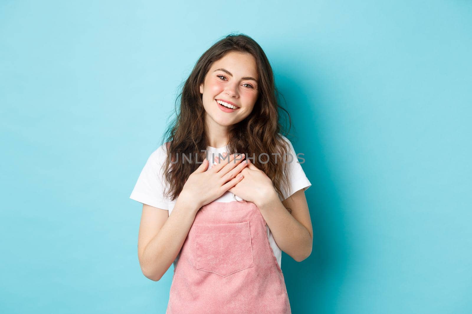 Portrait of grateful smiling young woman with curly hairstyle, summer make up, holding hands on heart and looking heartfelt, express gratitude, thanking you, blue background.