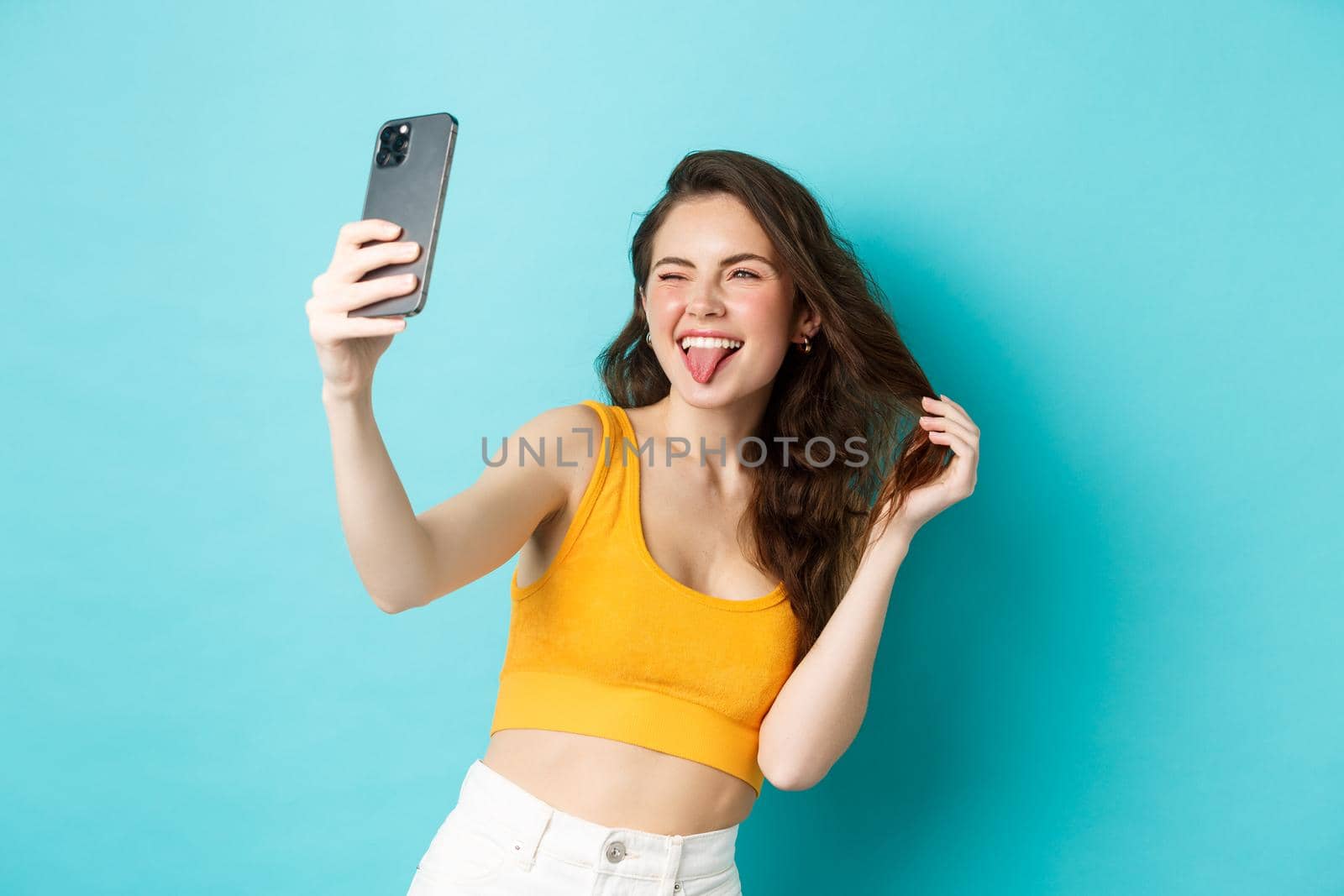 Technology and lifestyle concept. Happy young woman making silly faces while taking selfie on smartphone app with filters, standing against blue background.