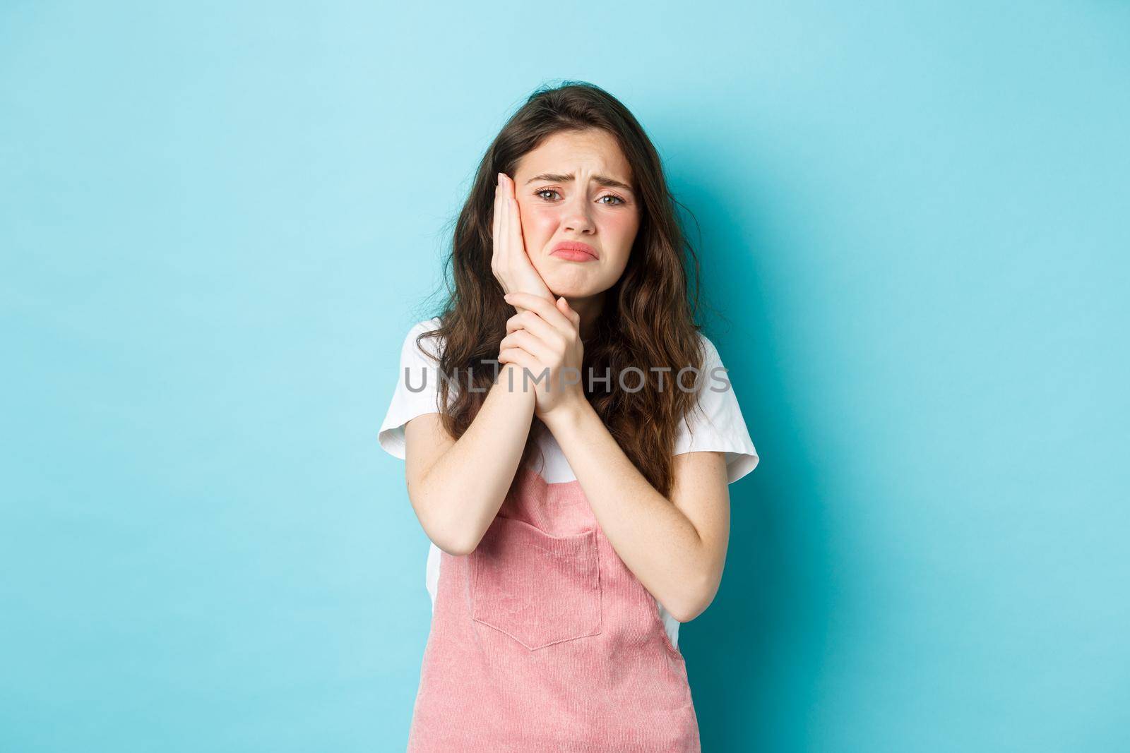 Image of frowning upset girl touching swallen cheek, having toothache, complaining on tooth decay, need to see dentist, standing against blue background by Benzoix