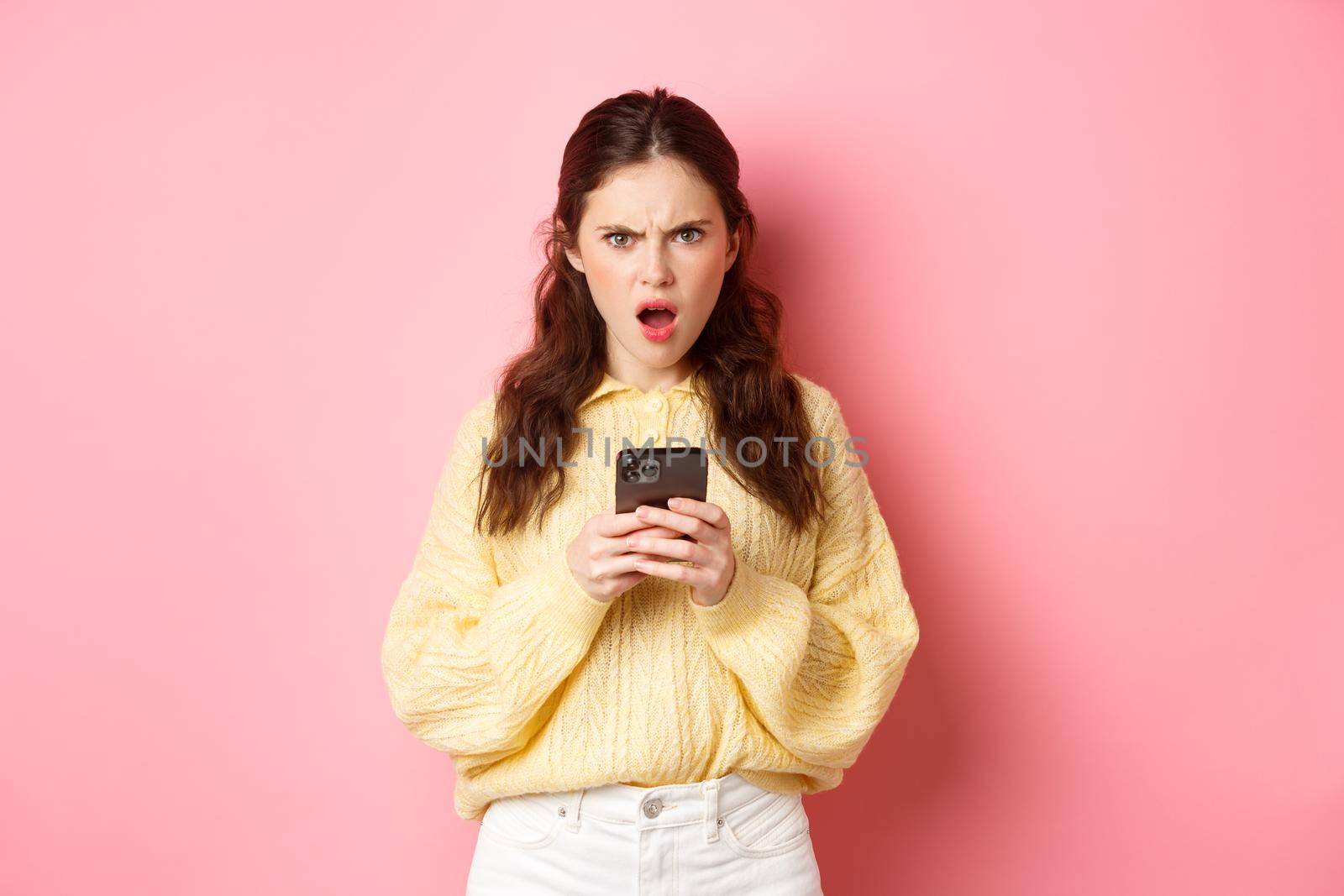 Confused and angry girlfriend reading strange message on phone, staring at camera frustrated and questioned, standing against pink background by Benzoix