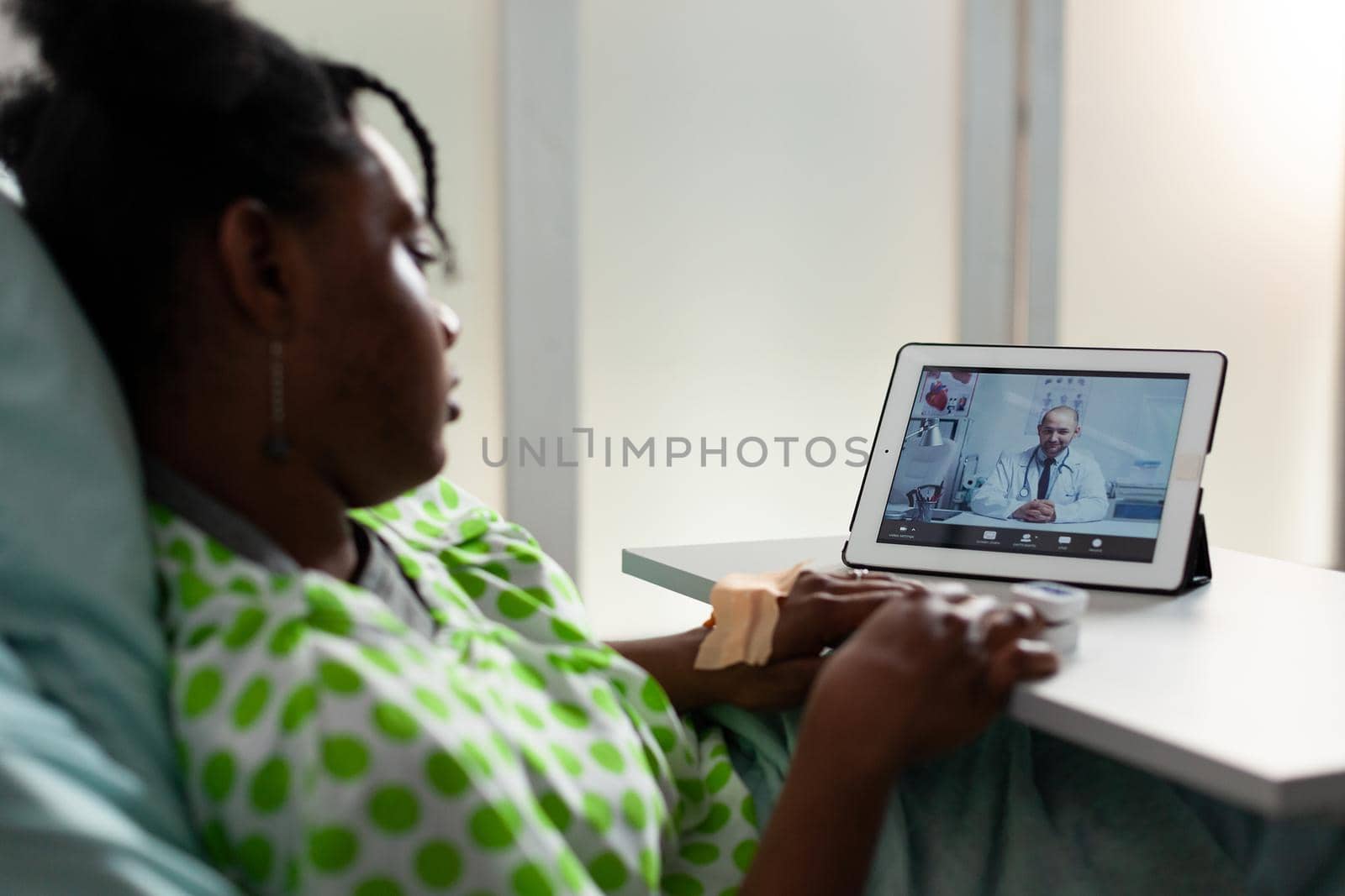 Young ill patient using tablet for telemedicine video call with doctor in hospital ward bed. Black person talking to specialist on online conference technology with digital gadget