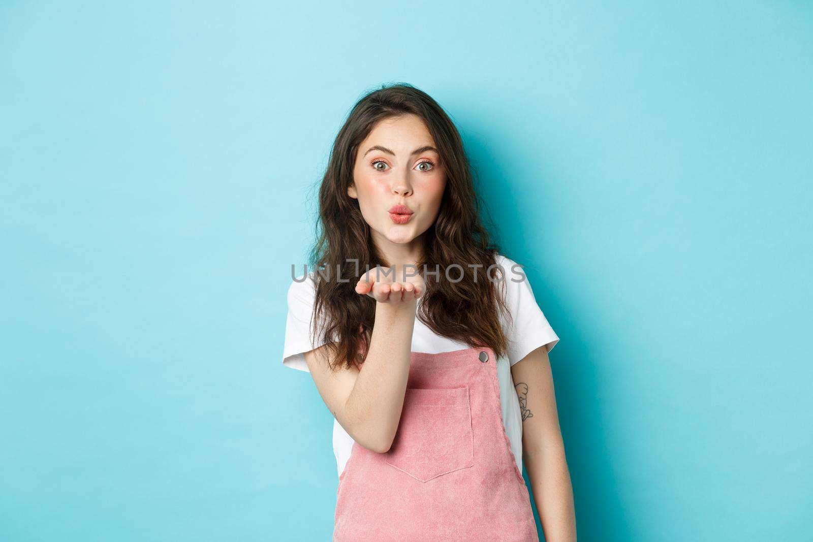 Beautiful girl blowing air kiss with romantic face, holding palm near lips and sending mwah at you, standing in summer outfit against blue background.