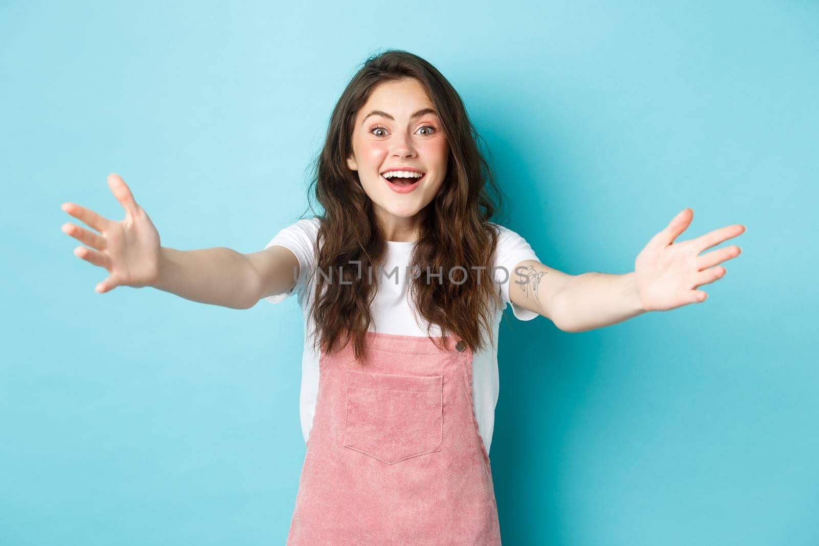 Image of happy young woman with curly hair and cute summer blush make up, stretch out hands to hug or hold someone, reaching for embraces, welcome you, blue background.