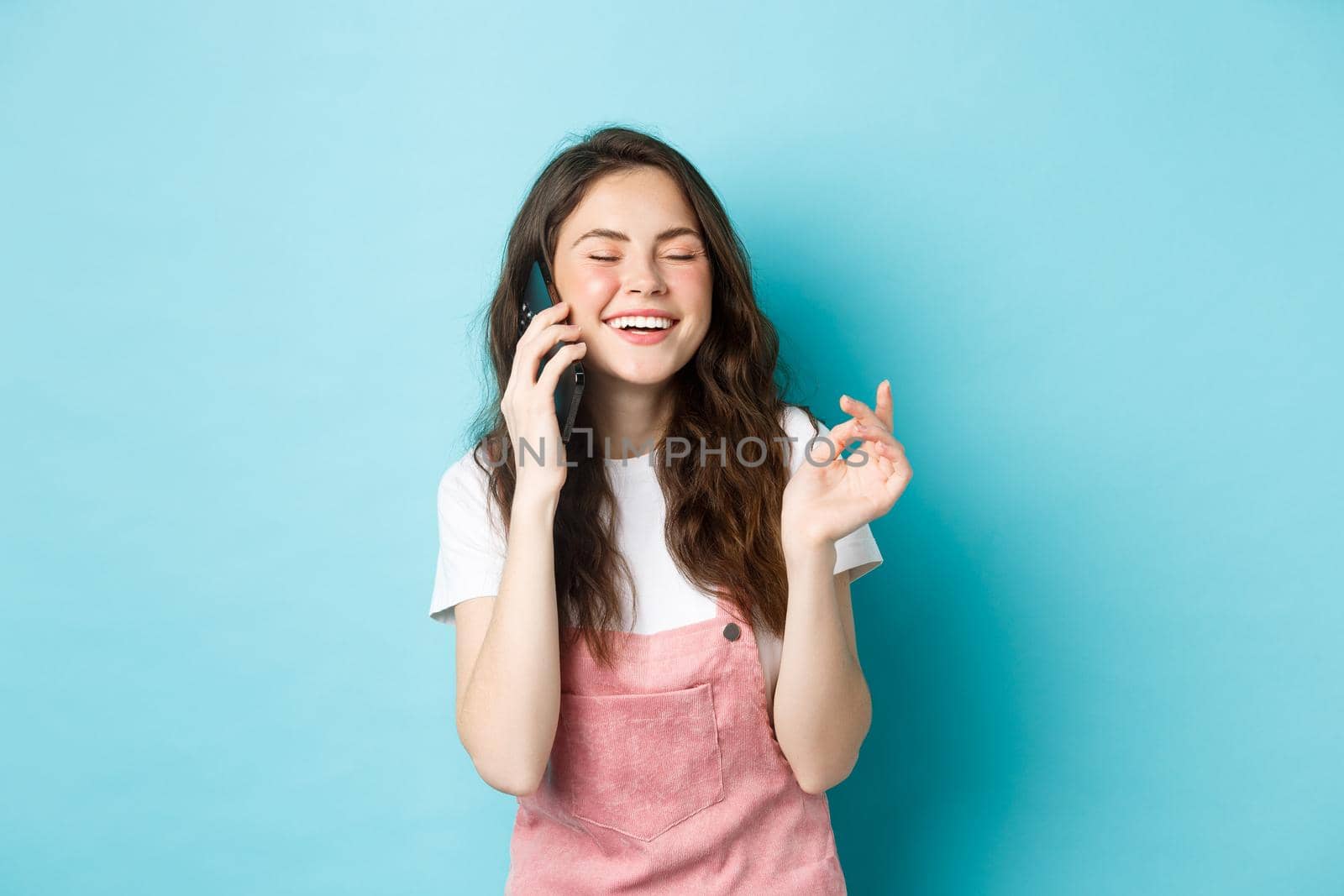 Portrait of cute and stylish girl laughing while chatting on phone, holding smartphone near ear and smiling happy, having casual conversation, standing over blue background.