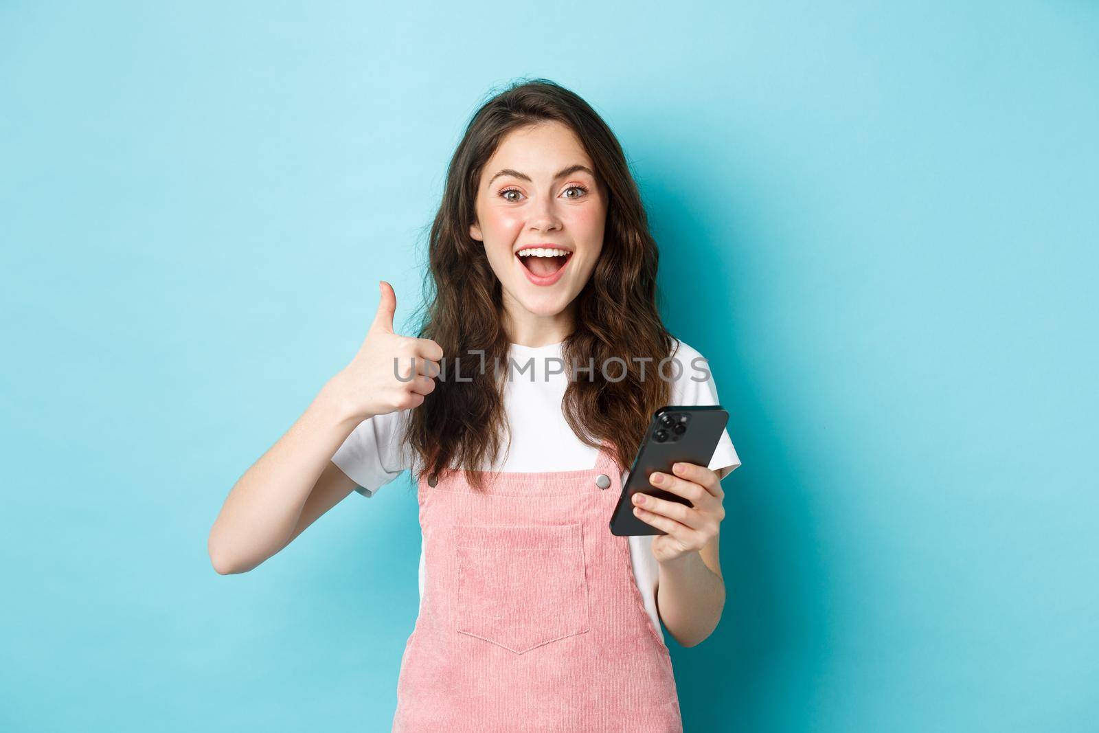 Portrait of excited cute girl say yes, smiling and nod in approval, holding smartphone while showing thumb up, like and praise good online offer, standing against blue background.