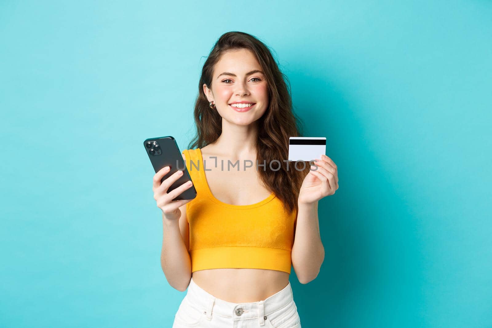 Online shopping. Beautiful woman getting ready for summer vacation, booking tickets with credit card and smartphone app, standing over blue background.