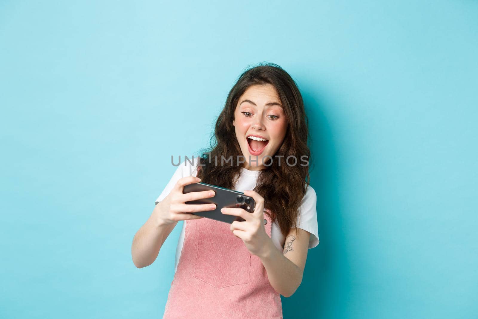 Portrait of excited young woman playing mobile video game with both hands, smiling amused, looking at screen, standing over blue background.