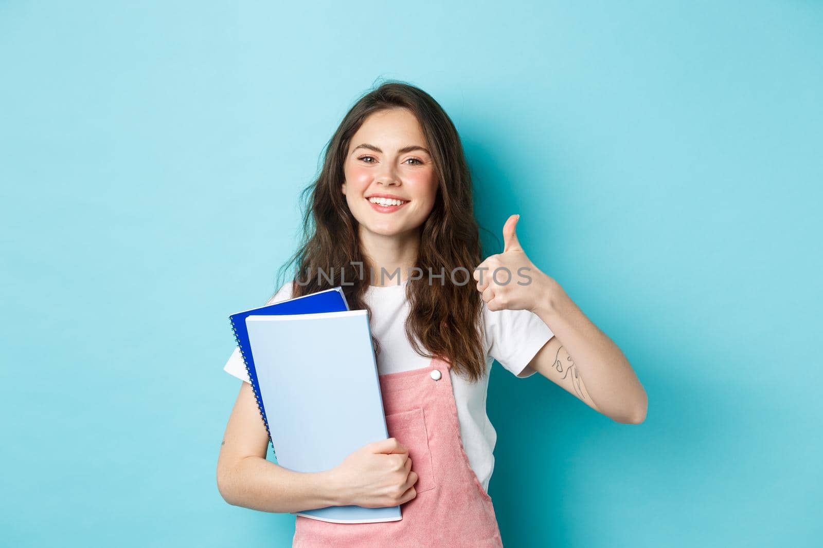 Young woman attend courses, girl student studying, holding notebooks and showing thumb up in approval, recommending company, standing over blue background.