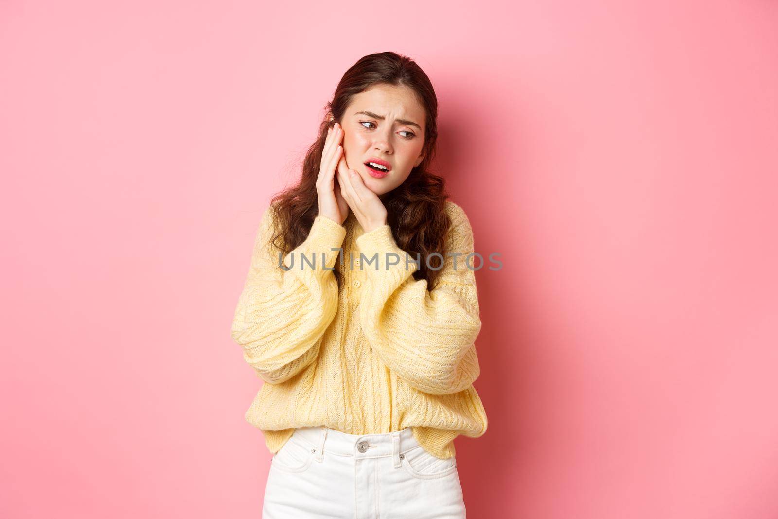 Young woman having toothache, touching swallen cheek and grimacing from pain, need to see dentist, stomatology appointment, standing against pink background.
