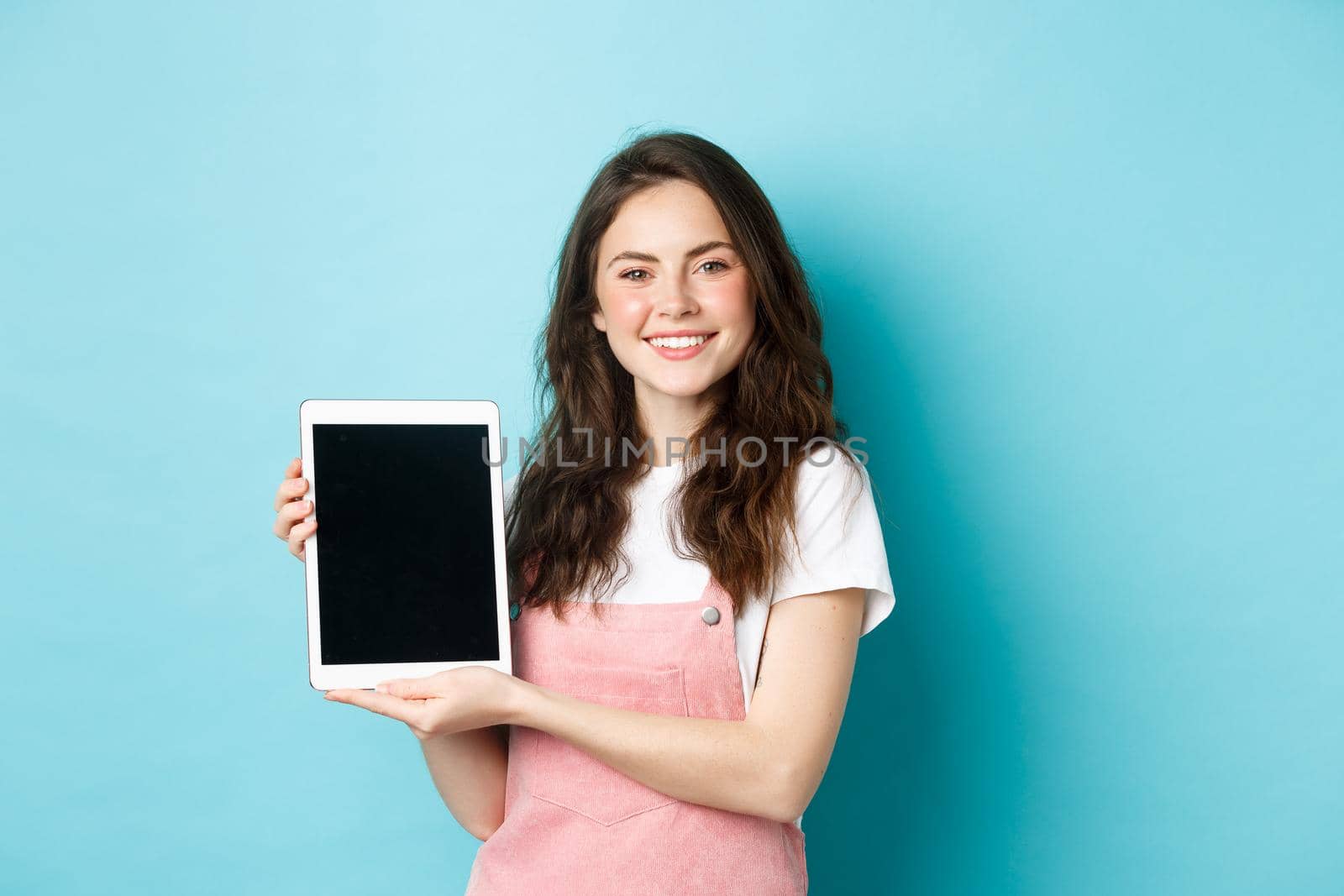 Portrait of smiling beautiful woman demonstrate blank tablet screen, showing promo on device screen, standing against blue background.