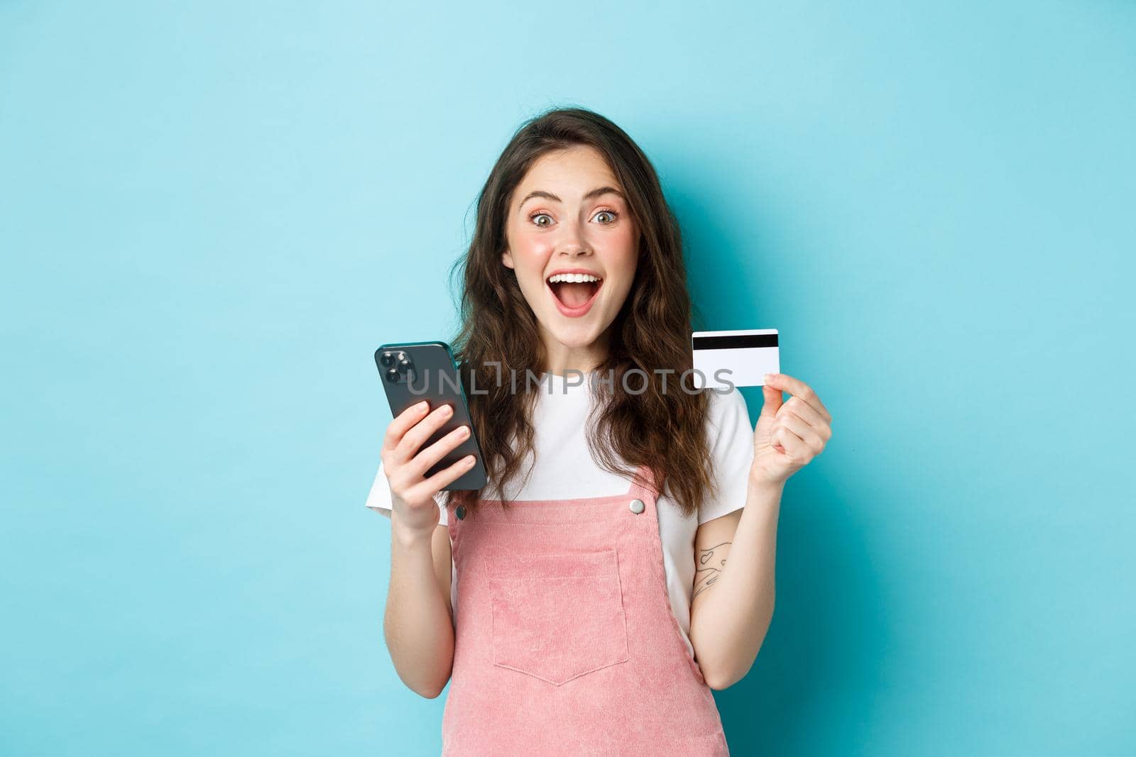 Surprised and happy young woman showing plastic credit card and smartphone, looking amazed at camera, shopping online with spring discounts, blue background by Benzoix