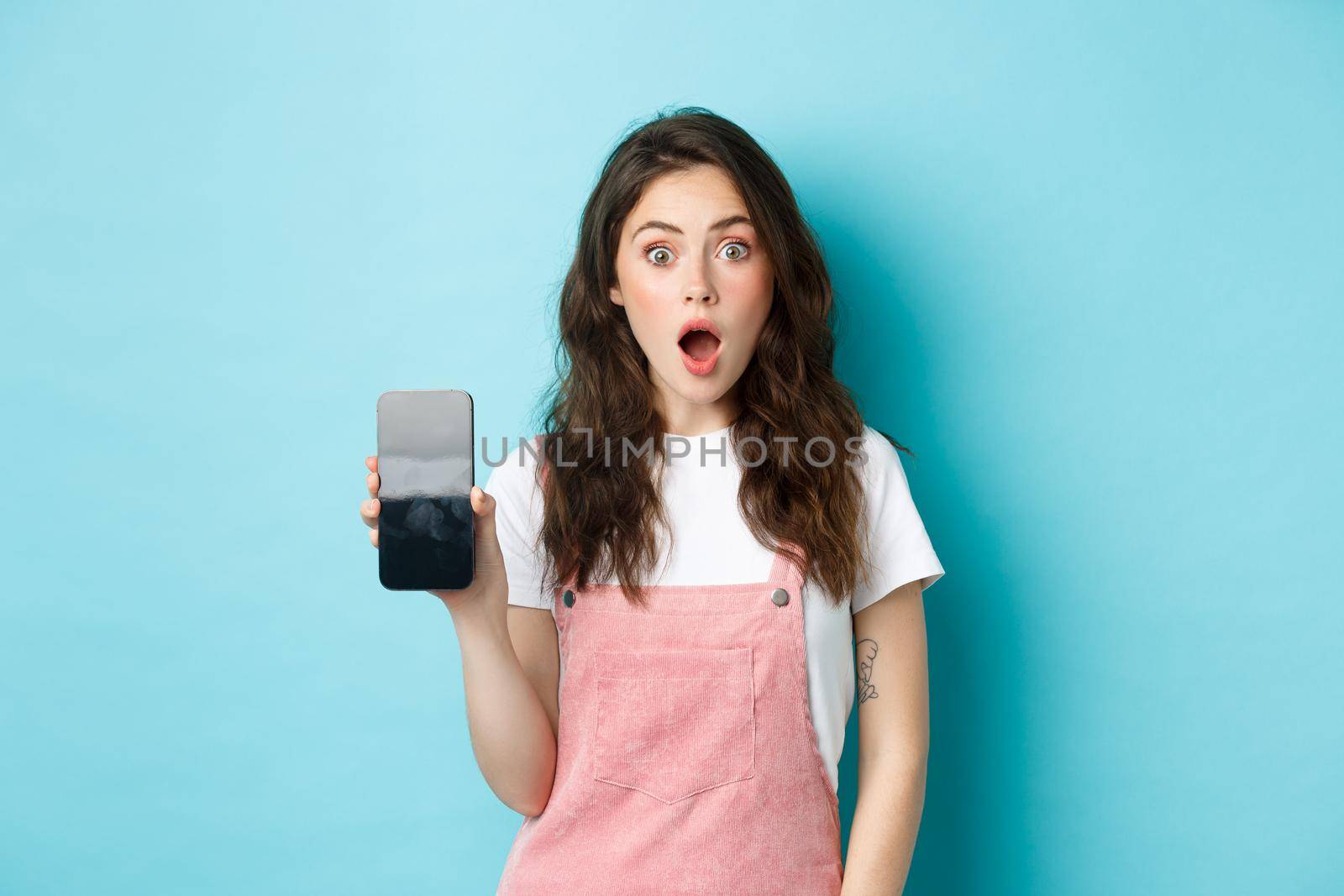 Shocked beautiful girl drop jaw and show empty smartphone screen, demonstrate your app or online store on blank phone display, standing against blue background by Benzoix