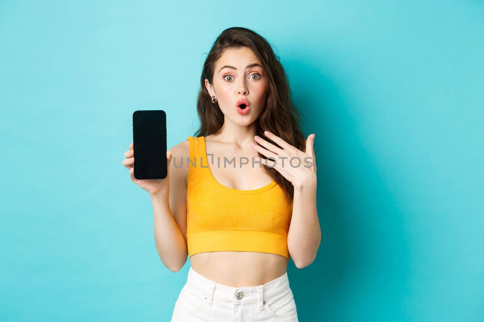 Portrait of surprised girl impressed with smartphone app, showing empty mobile screen and look amazed, standing over blue background.