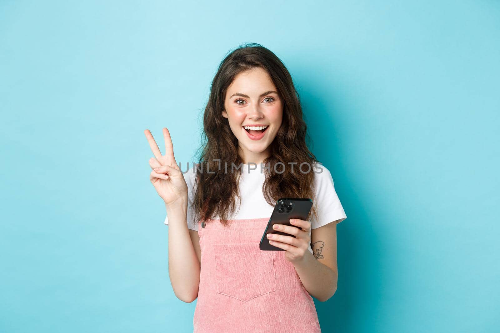 Online shopping. Positive smiling girl showing v-sign and looking happy at camera, using smartphone, holding mobile phone, standing against blue background.