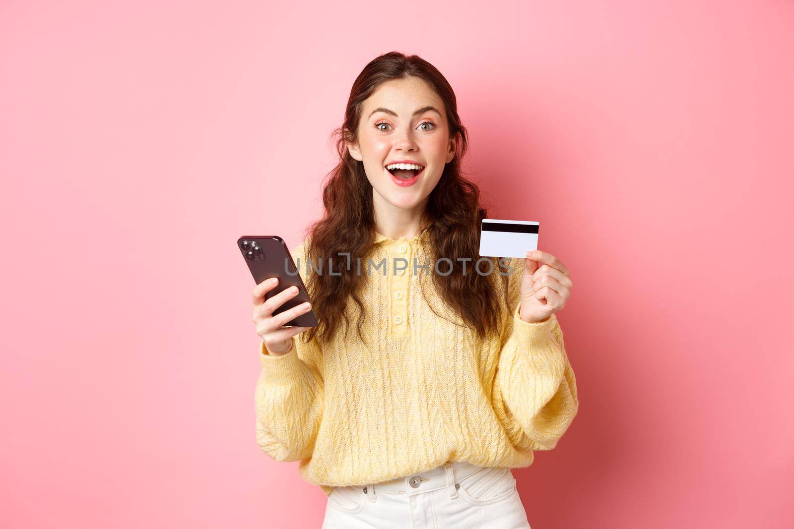 Technology and online shopping. Excited girl making order, paying online with plastic credit card, holding mobile phone and smiling at camera, pink background by Benzoix