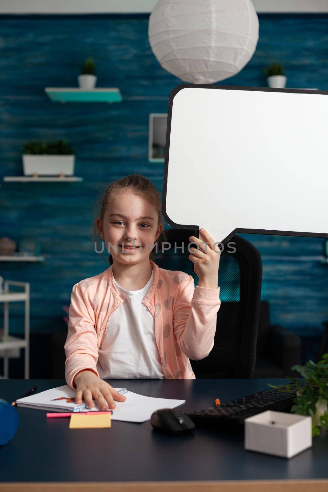 Portrait of smiling little schoolchild holding white board looking into camera during virtual school lesson in living room. Concept of home schooling, distance learning, online education