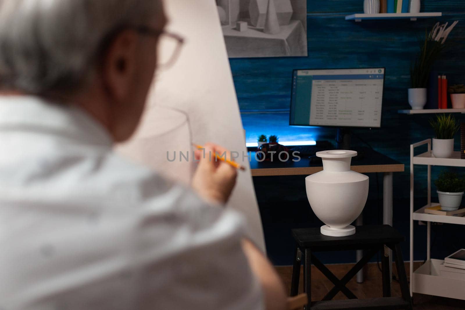 Caucasian artist looking at vase on desk and drawing on canvas at workplace. Senior man with artistic hobby recreating professional masterpiece using art intruments and tools.