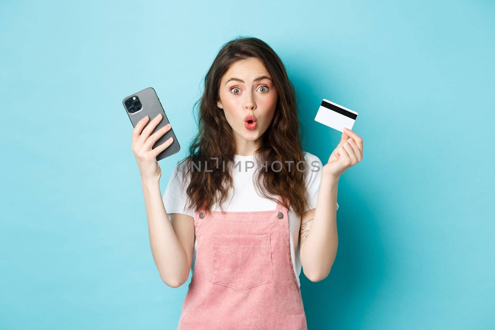 Surprised young woman say wow, stare excited at camera, holding mobile phone and plastic credit card, standing over blue background.