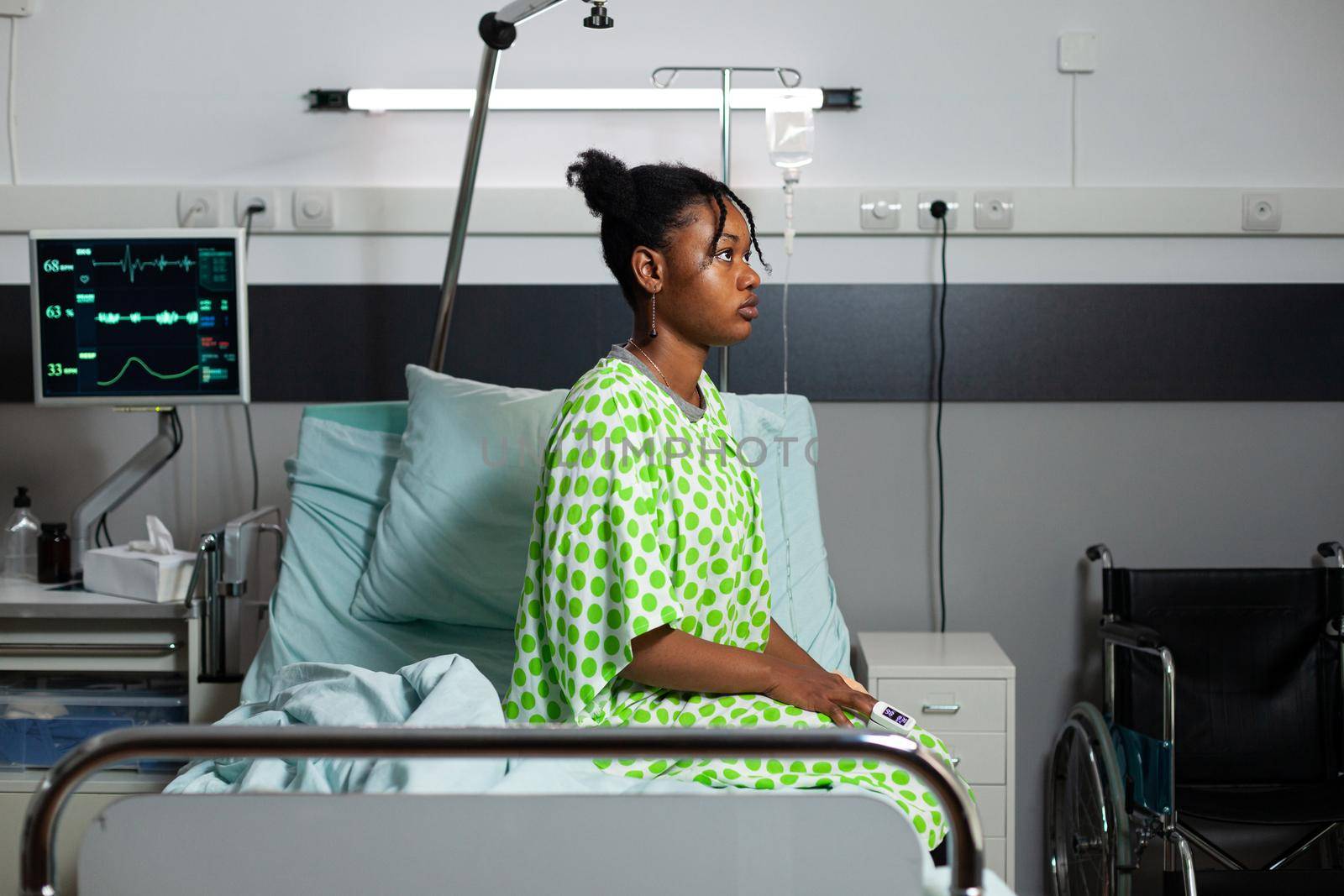 Young african american person sitting on hospital ward bed with fever, sickness, disease. Sick teenager with oximeter on hand, medical equipment and heart rate monitor for recovery