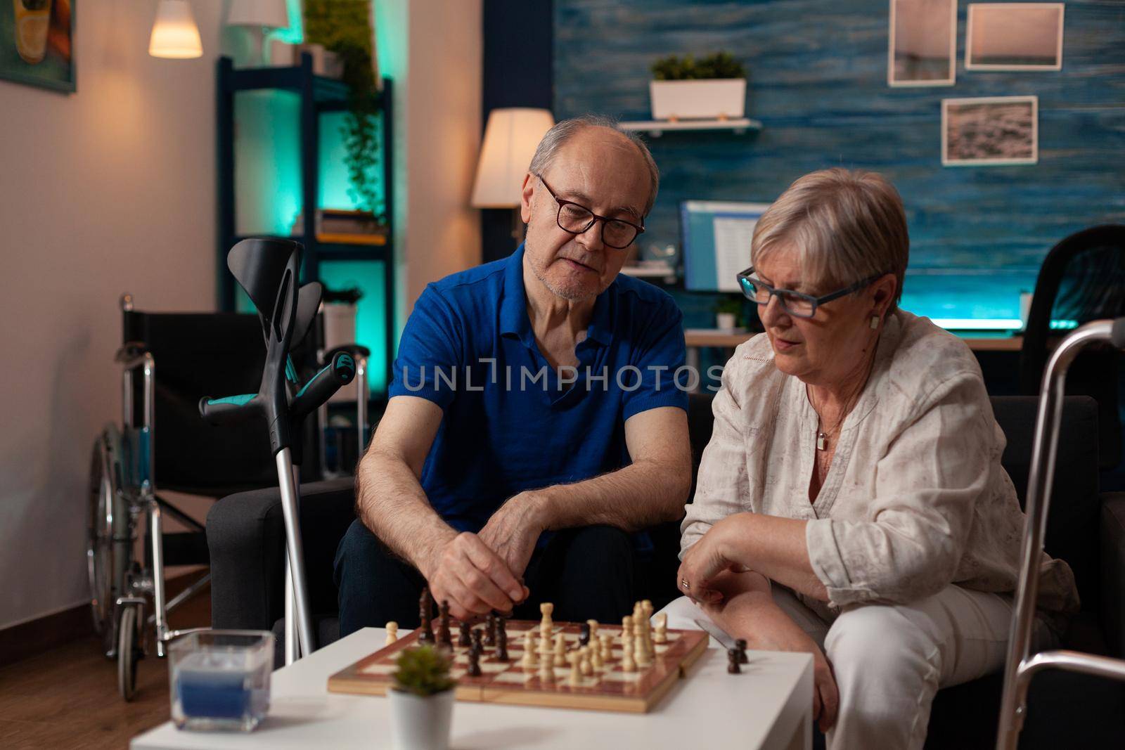 Senior people enjoying retirement with chess game on coffee table in living room. Elderly couple sitting on couch with chessboard playing together for entertainment and fun at home