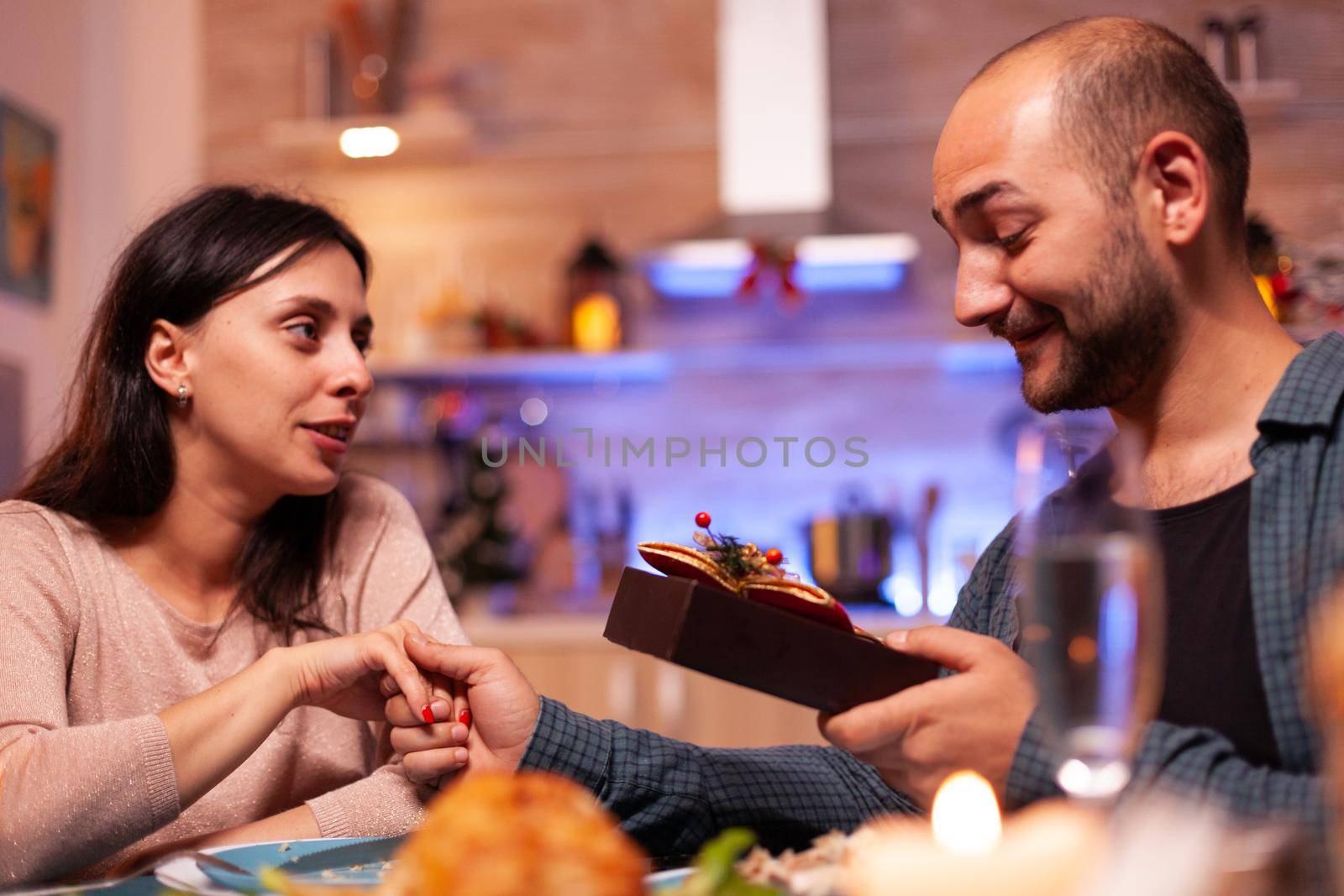 Happy couple enjoying christmas holiday surprising with xmas present with ribbon on it sitting at table in x-mas decorated kitchen. Cheerful family celebrating winter holiday spending time together