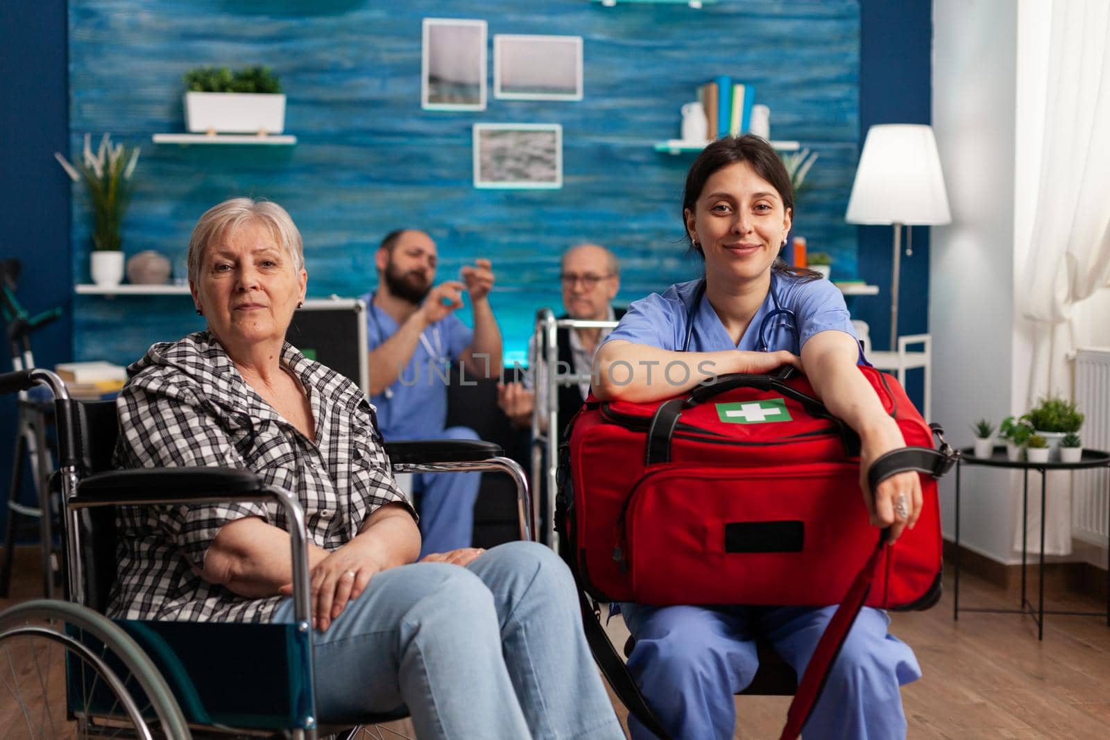 Portrait of support nurse worker holding medicine kit bag sitting beside disabled senior patient looking into camera during medical therapy. Healthcare assistance. Social services nursing at home