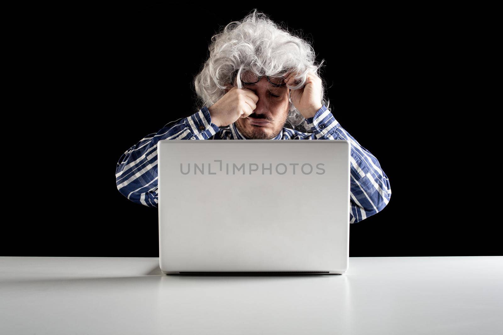 Portrait of a senior man rubbing his tired eyes sitting in front of the laptop computer on a white table. Black background