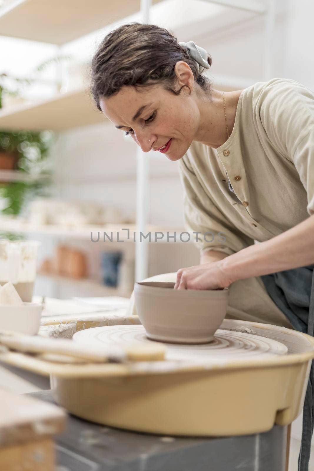 Lady siting on bench with pottery wheel and making clay pot by Antonelli