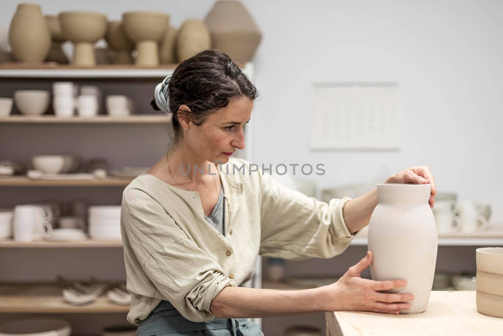 Young woman holding pottery instrument for scraping, smoothing, shaping and sculpting. Lady siting on bench with pottery wheel and making clay pot