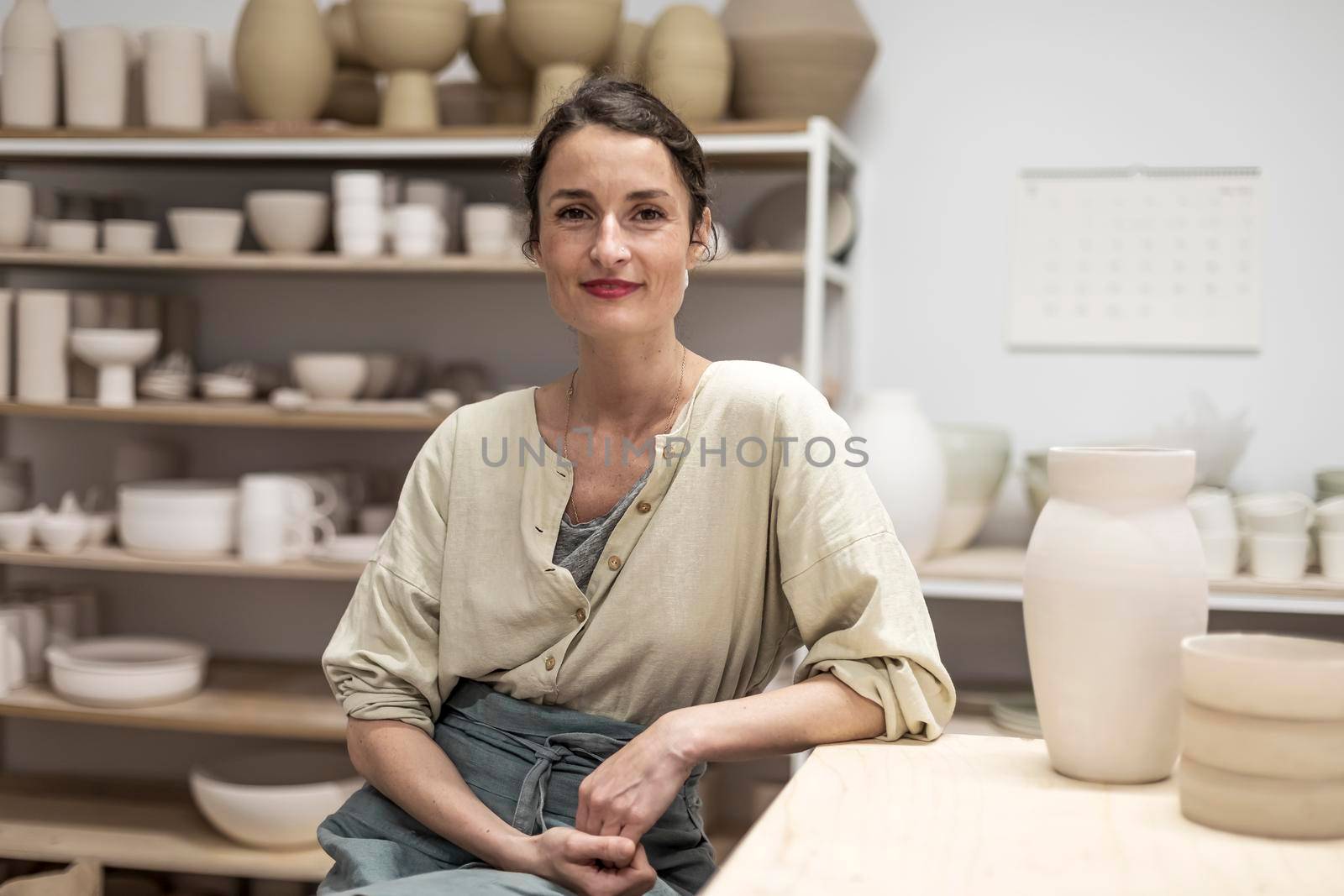 Beautiful happy craft woman wearing apron looking at camera and smiling while sitting in her art studio or craft pottery shop