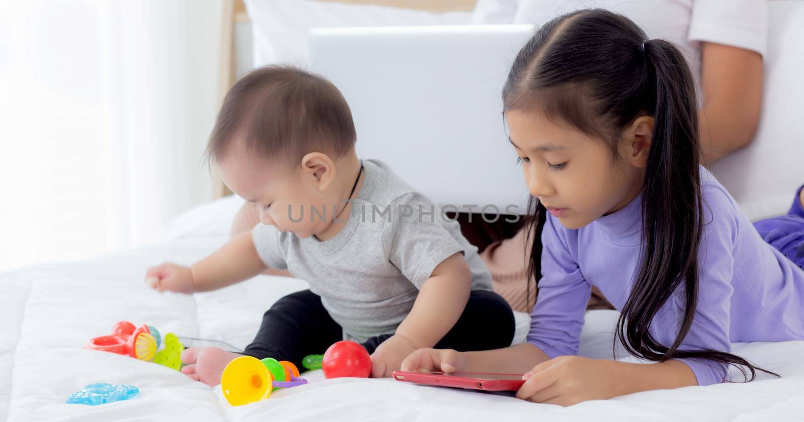 Young asian mother working at home online to internet with laptop computer and daughter looking tablet on bed at bedroom, business woman is freelance and children learning, new normal, family concept. by nnudoo