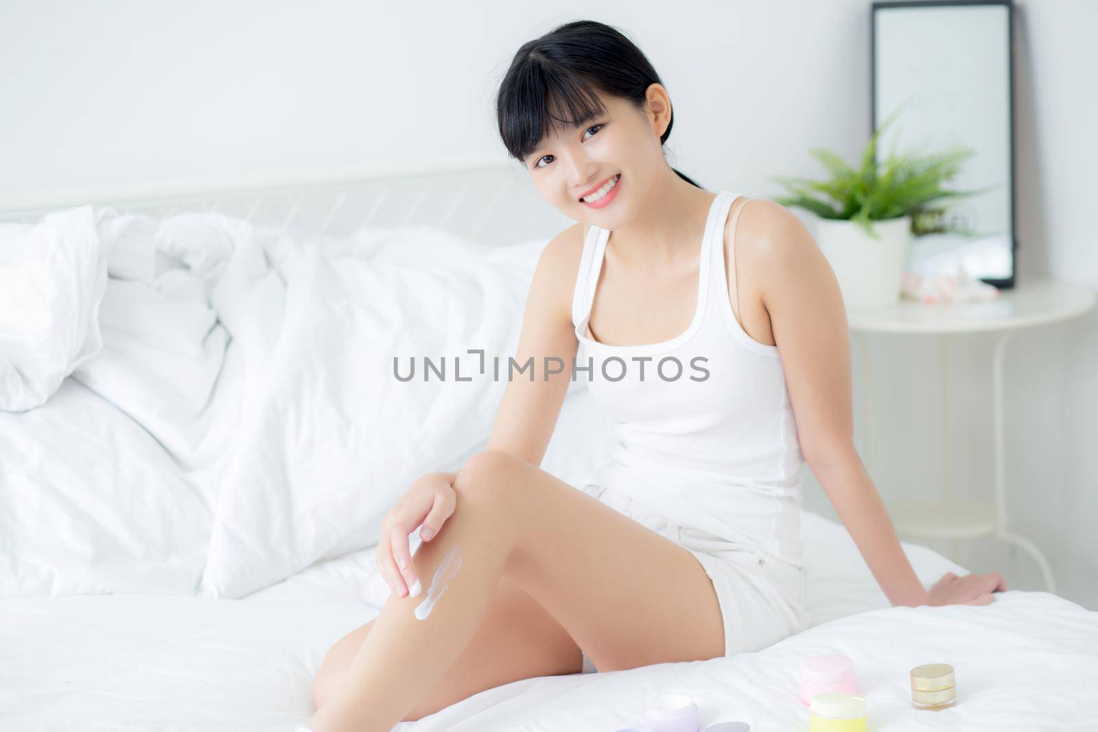Beautiful young asian woman applying cream or lotion on skin legs with moisturizer having cartridge on bed, beauty asia girl applying skincare touch legs with cosmetic makeup, skin care concept. by nnudoo