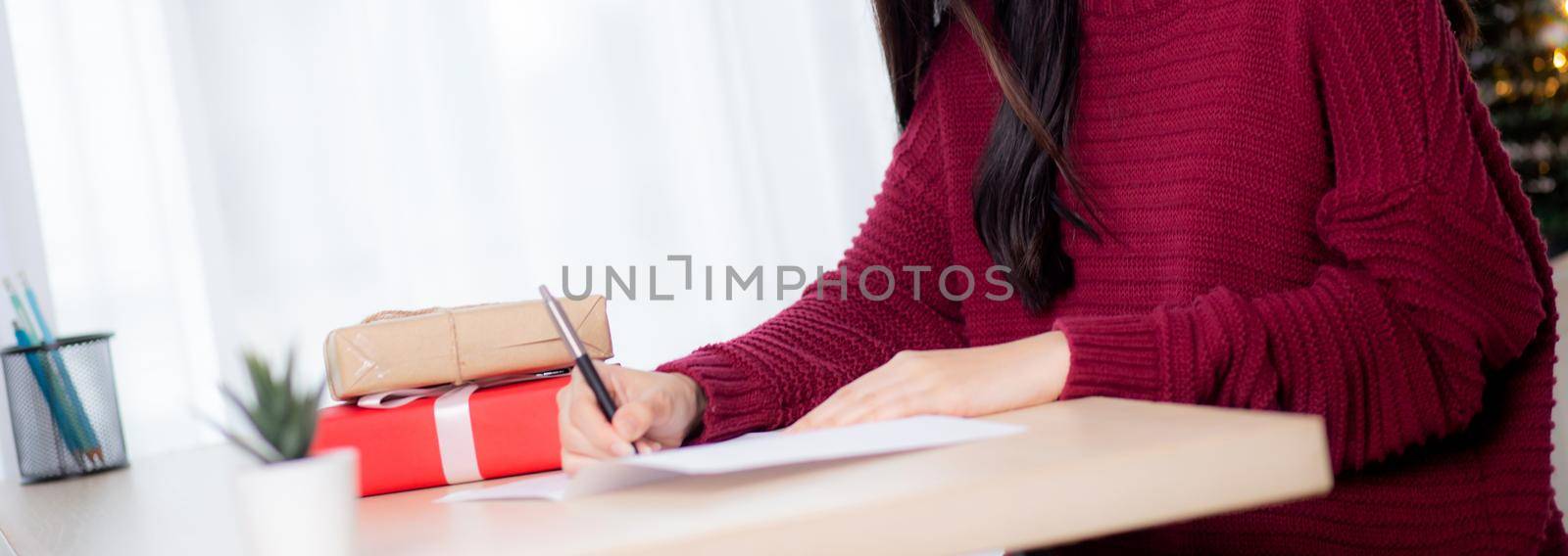 Closeup hand of young asian woman writing postcard in Christmas day at home, eve and celebrate, female writing message on greeting card with giving gift box in holiday, congratulation and celebration. by nnudoo