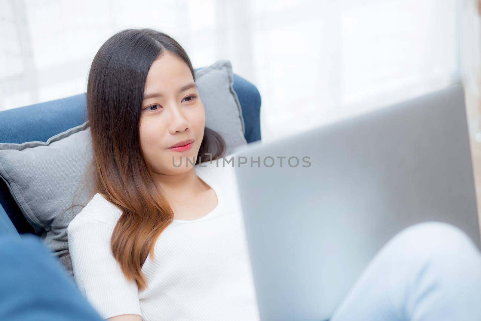 Young asian business woman smile and work from home with laptop computer online to internet on sofa in living room, freelance girl using notebook on couch with comfort, new normal, lifestyle concept. by nnudoo
