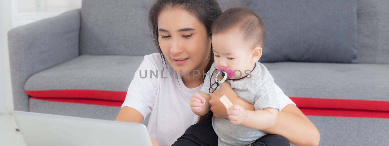 Young asian mother working on laptop computer while care daughter girl at home, mom and baby girl sitting using notebook video call, parent and bonding, indoors, family and business concept. by nnudoo
