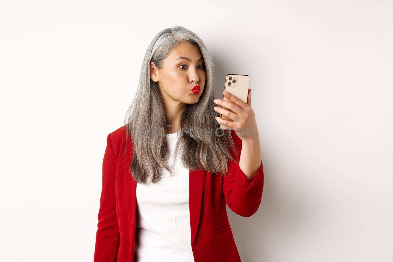 Silly and funny asian senior woman pouting, taking selfie on smartphone, standing over white background.