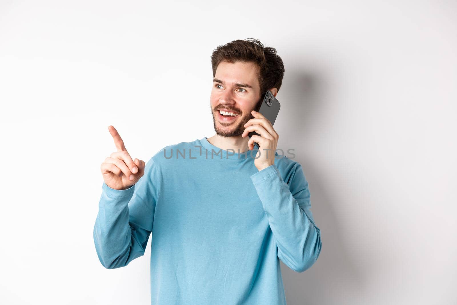 Technology concept. Young man talking on mobile phone and pointing left, having call on smartphone, standing over white background by Benzoix