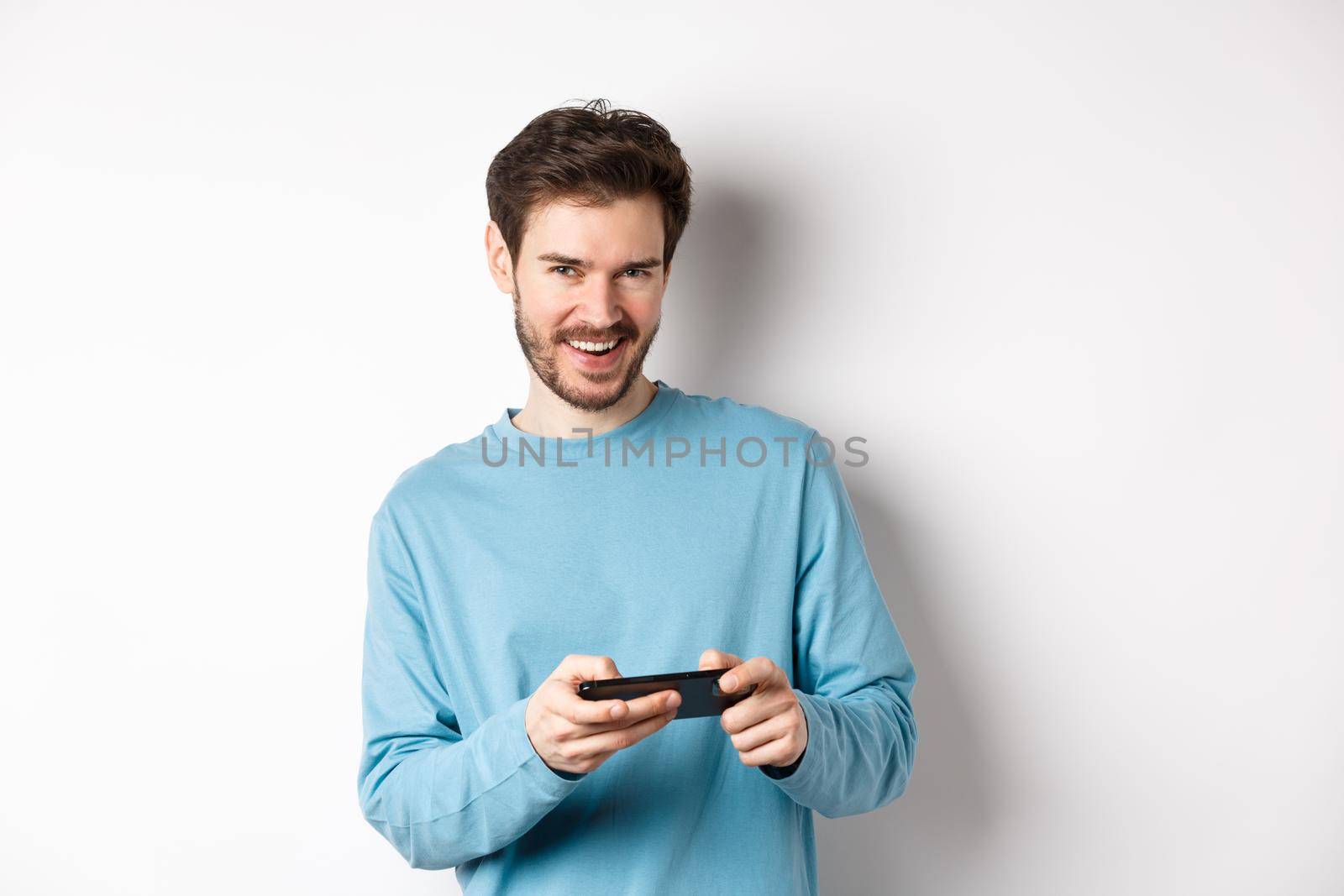 Image of handsome young man playing mobile video game, holding smartphone horizontally and smiling at camera pleased, standing over white background by Benzoix