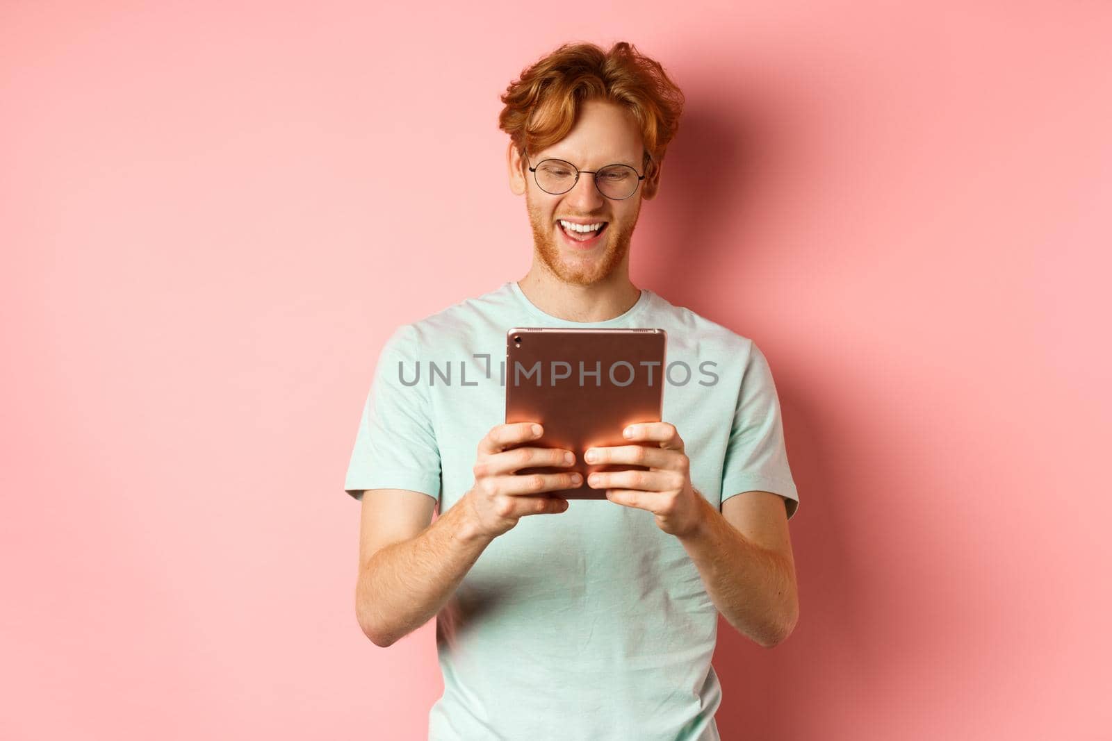 Happy guy with red hair and beard using digital tablet, reading screen and smiling, standing over pink background.